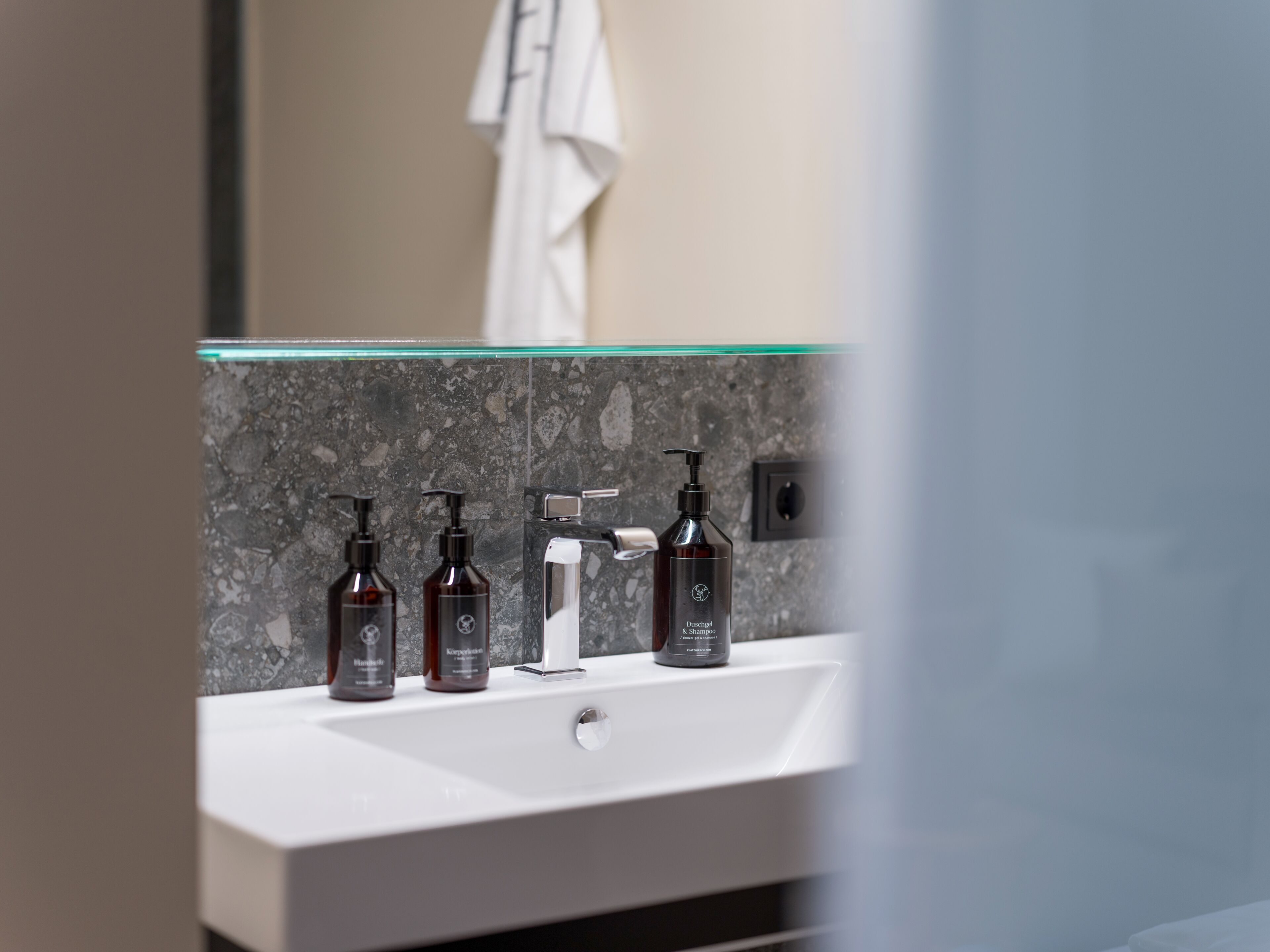 Un baño con un lavamanos blanco, una mampara de cristal y tres botellas marrón oscuro de champú.