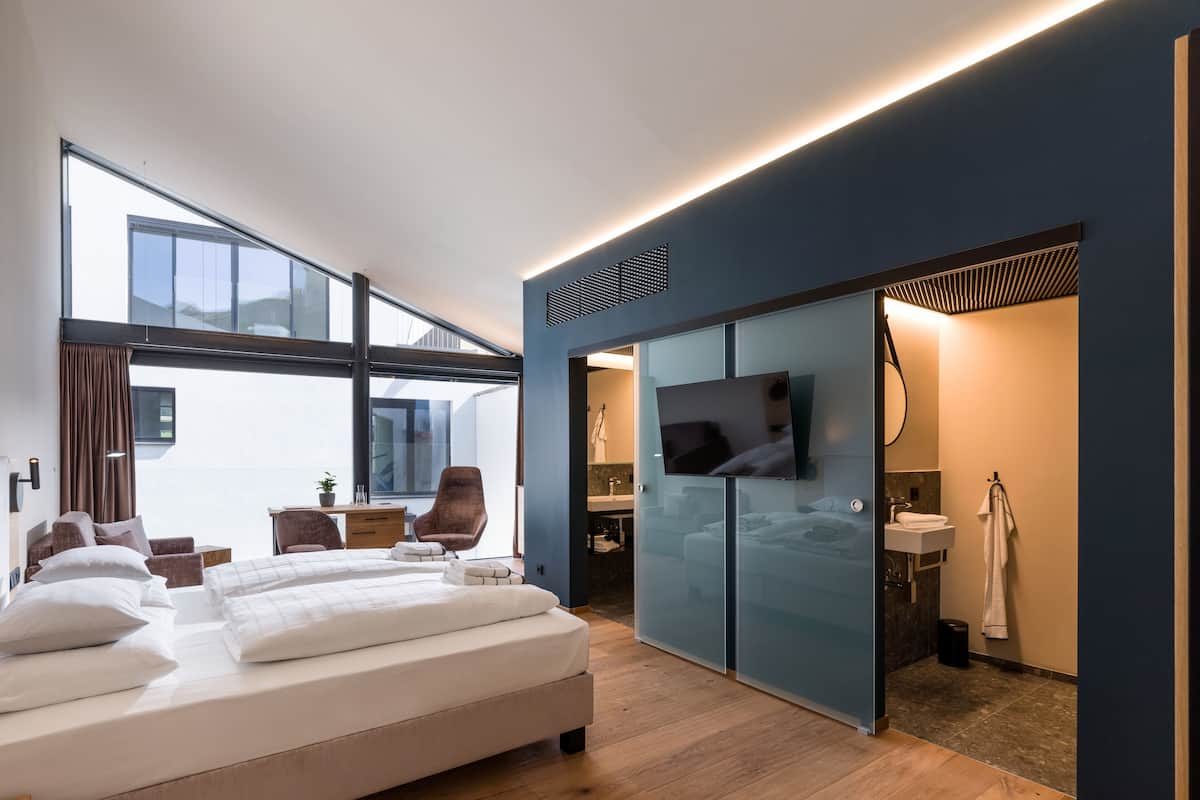 A modern hotel room with a large bed, a desk, a chair, a TV, and a bathroom with a mirror and sink.