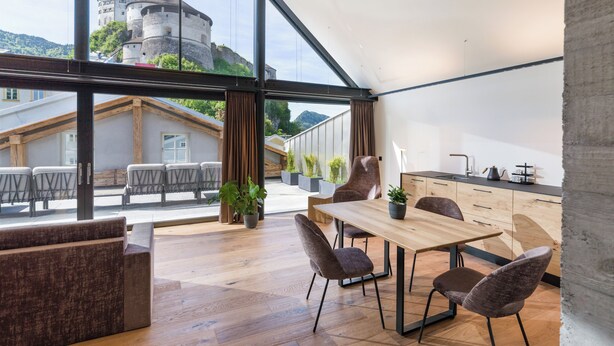 A modern interior with a wooden dining table, chairs, and a large window overlooking a castle. - Platzhirsch Kufstein (Kufstein)