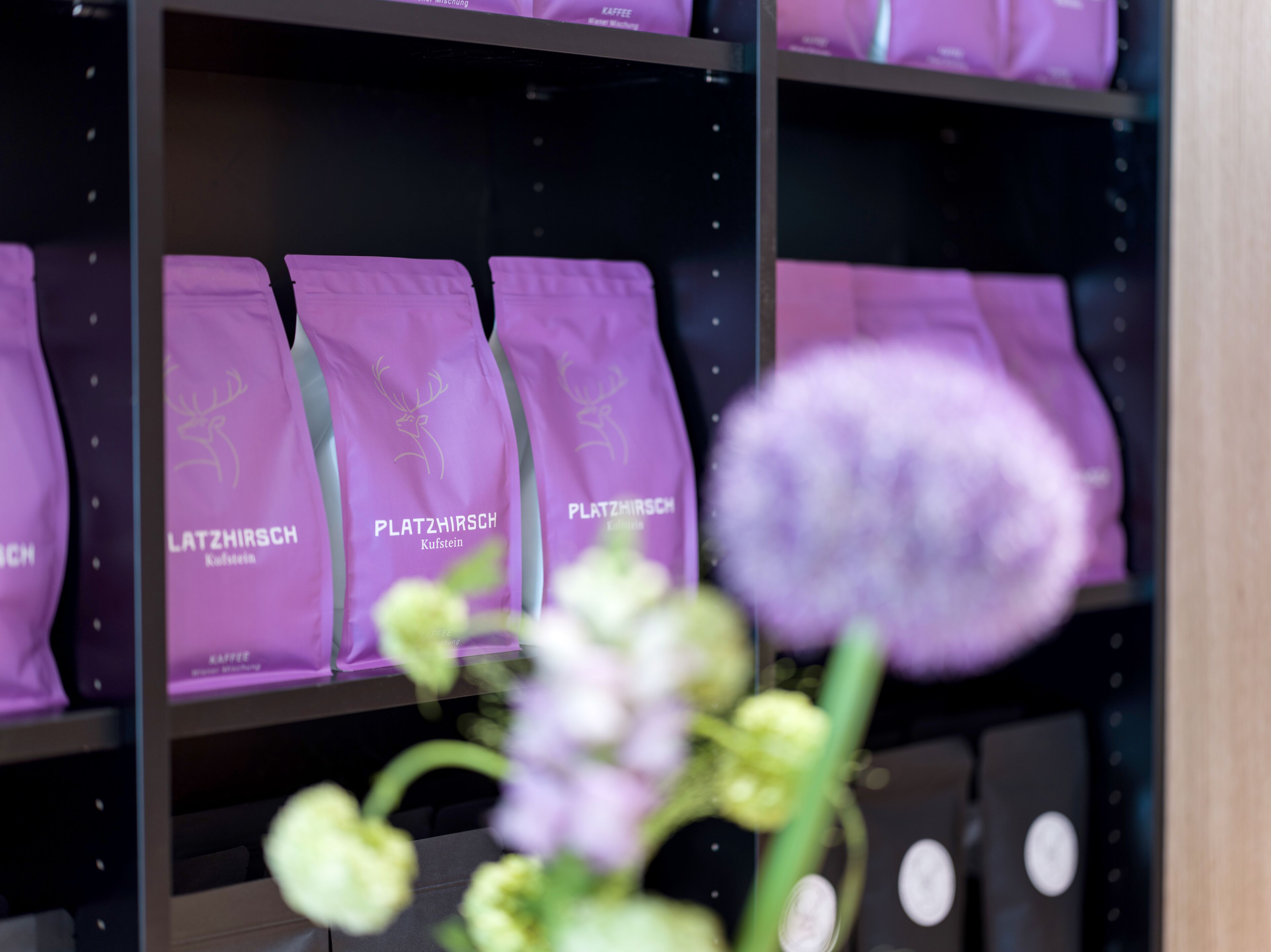 Estantes con bolsas moradas etiquetadas 'PLATZHIRSCH' y una flor borrosa en primer plano.