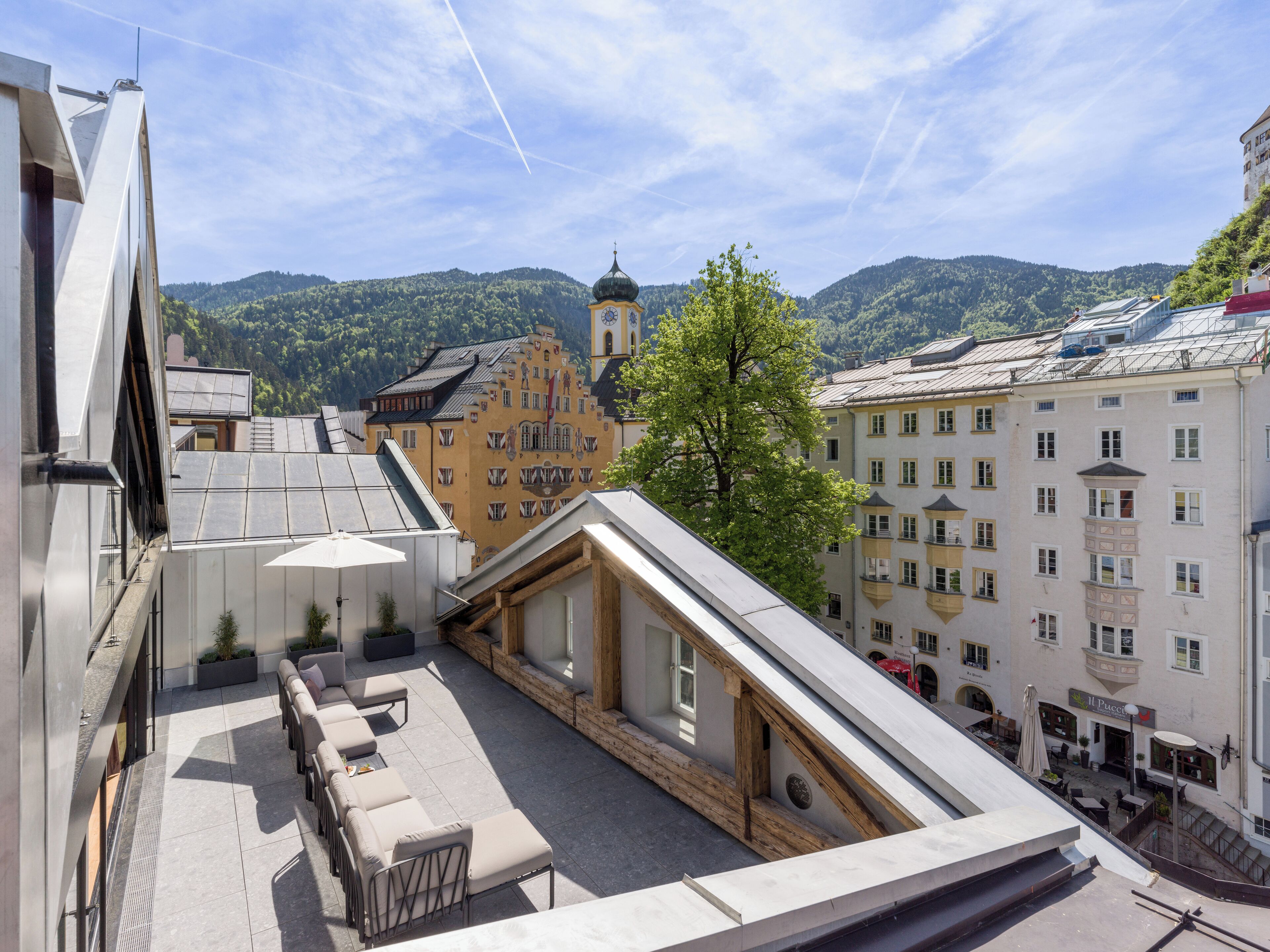 Una terraza en la azotea con mobiliario exterior, vistas a un edificio histórico y montañas al fondo.