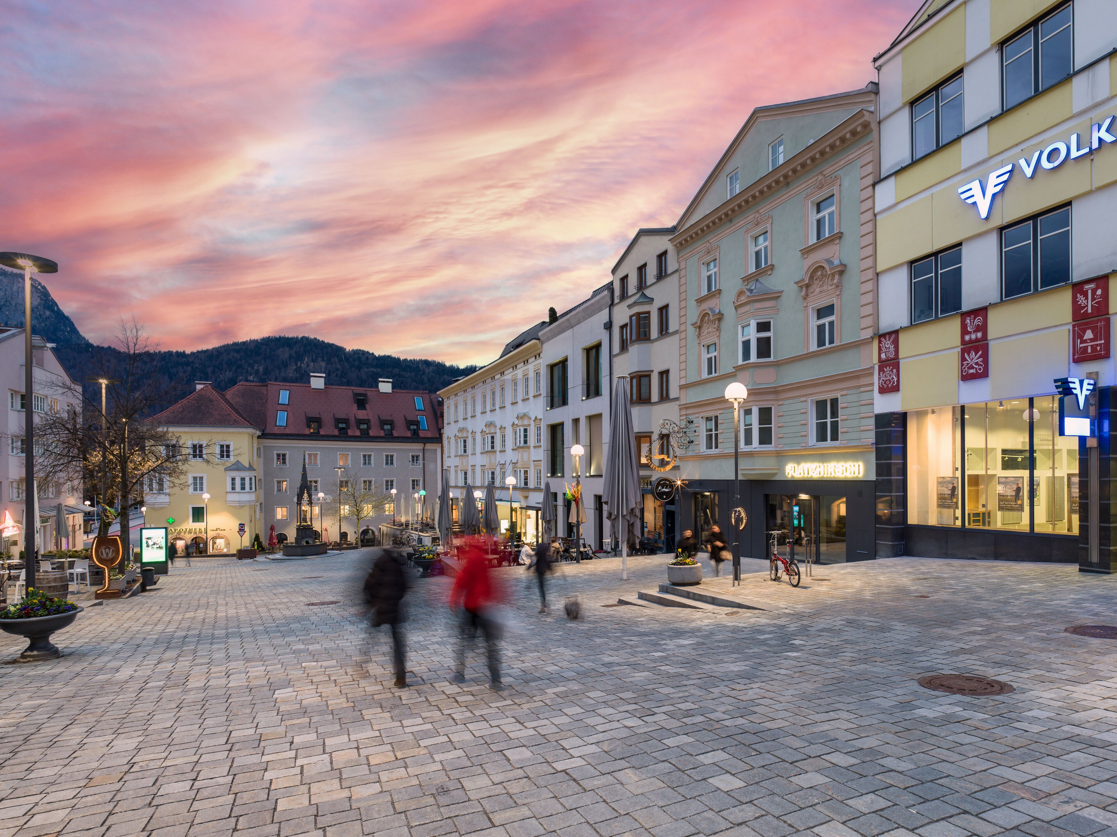 A cobblestone square with historic buildings, a fountain, and outdoor seating areas.