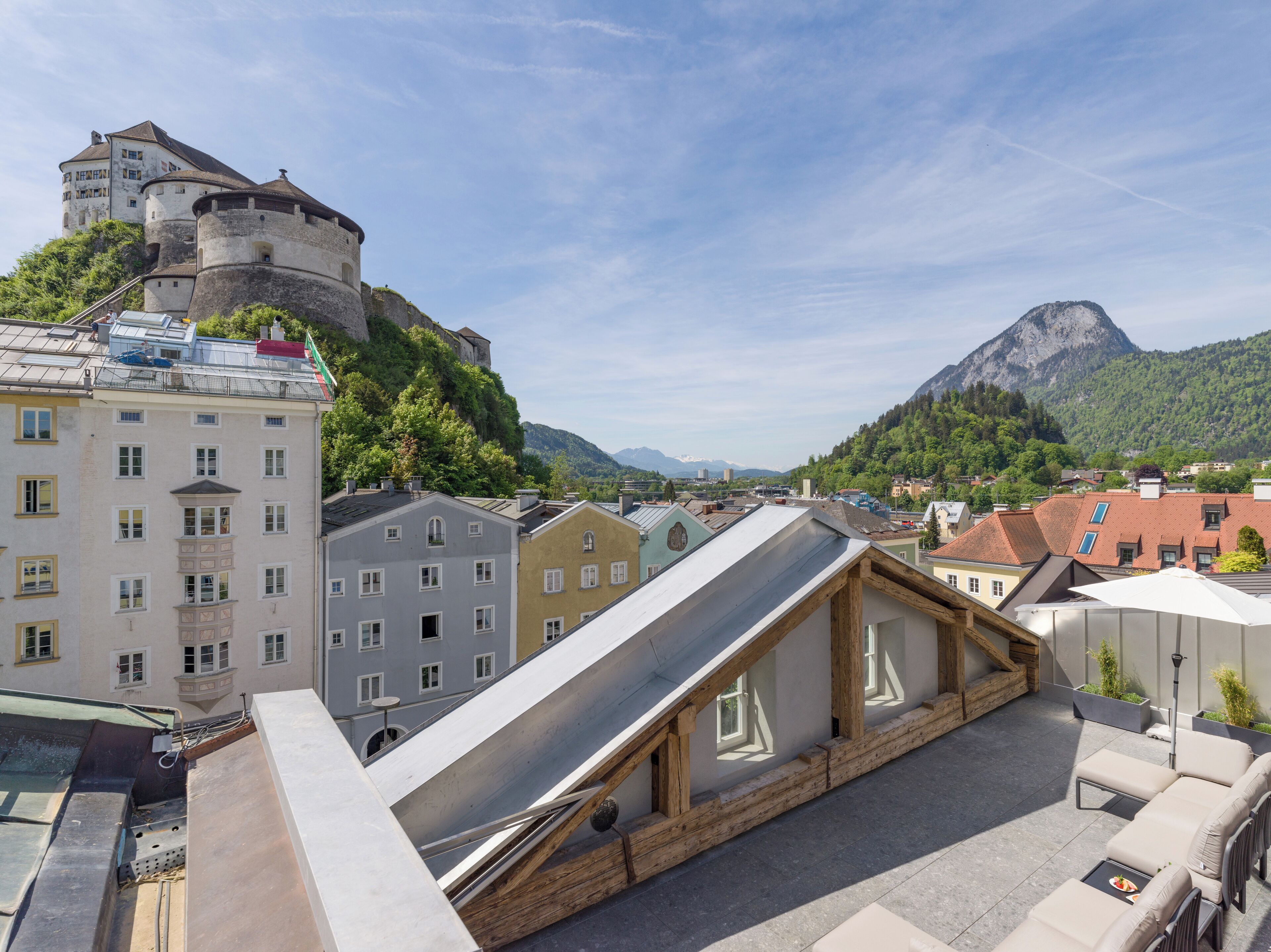A rooftop terrace with a modern metal roof, wooden beams, and a view of a historic castle and mountains.