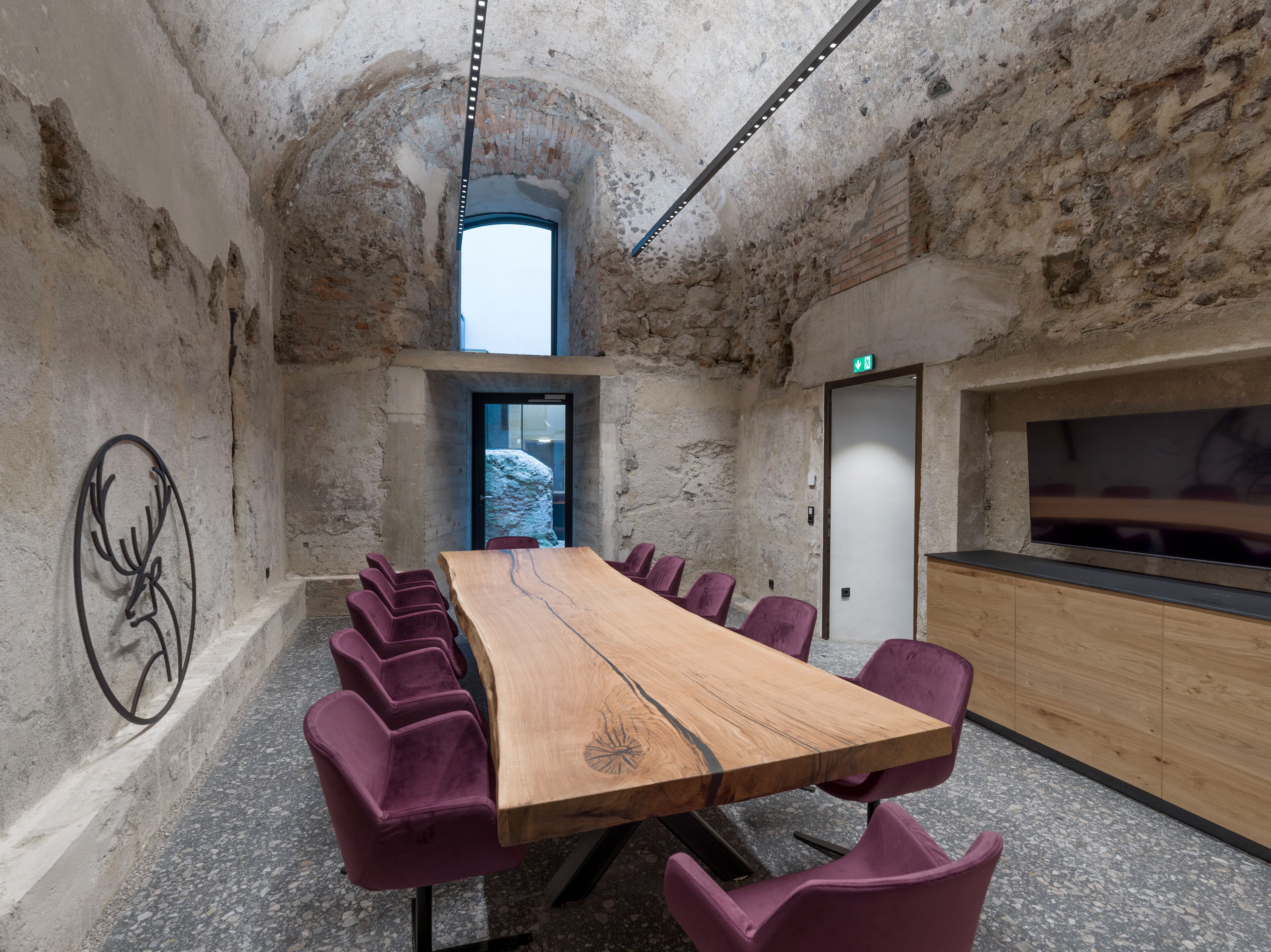 Uma sala de conferências com uma mesa de madeira comprida, cadeiras roxas, uma parede de pedra e uma televisão à direita.
