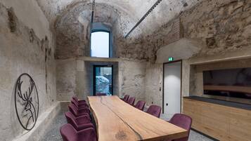 Uma sala de conferências com uma mesa de madeira comprida, cadeiras roxas, uma parede de pedra e uma televisão à direita.