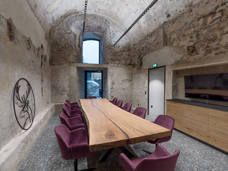 Uma sala de conferências com uma mesa de madeira comprida, cadeiras roxas, uma parede de pedra e uma televisão à direita.