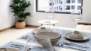 A dining table set with plates, bowls, and cutlery, a potted plant, and a window with a city view. - Bcons Garden Luxury Apartment (Ho Chi Minh City)