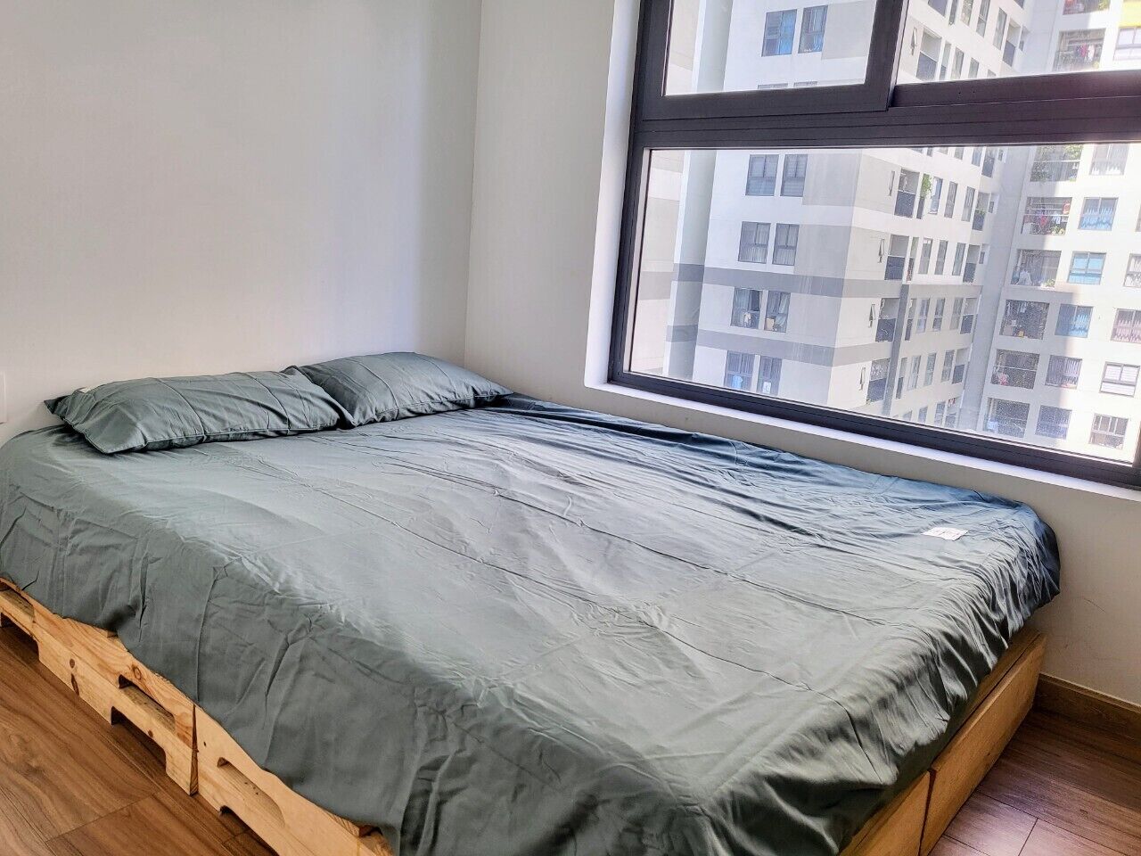 Een eenpersoonsbed met een grijze matras en kussens, een houten frame en een raam met uitzicht op een gebouw.