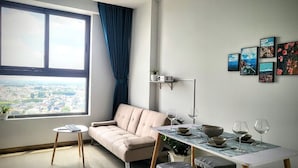 A modern living room with a sofa, a dining table set for two, a window with a view of the city, and a wall with framed pictures. - Bcons Garden Luxury Apartment (Ho Chi Minh City)
