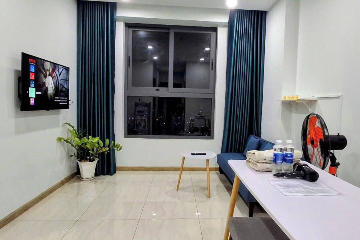 Une pièce dotée d’une grande fenêtre, d’un téléviseur à écran plat, d’une petite table avec un ventilateur, d’une chaise et d’une plante en pot.