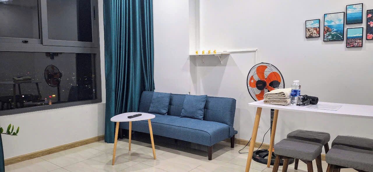 A room with a blue sofa, a small round table, a white table with a fan, and a window with a curtain.