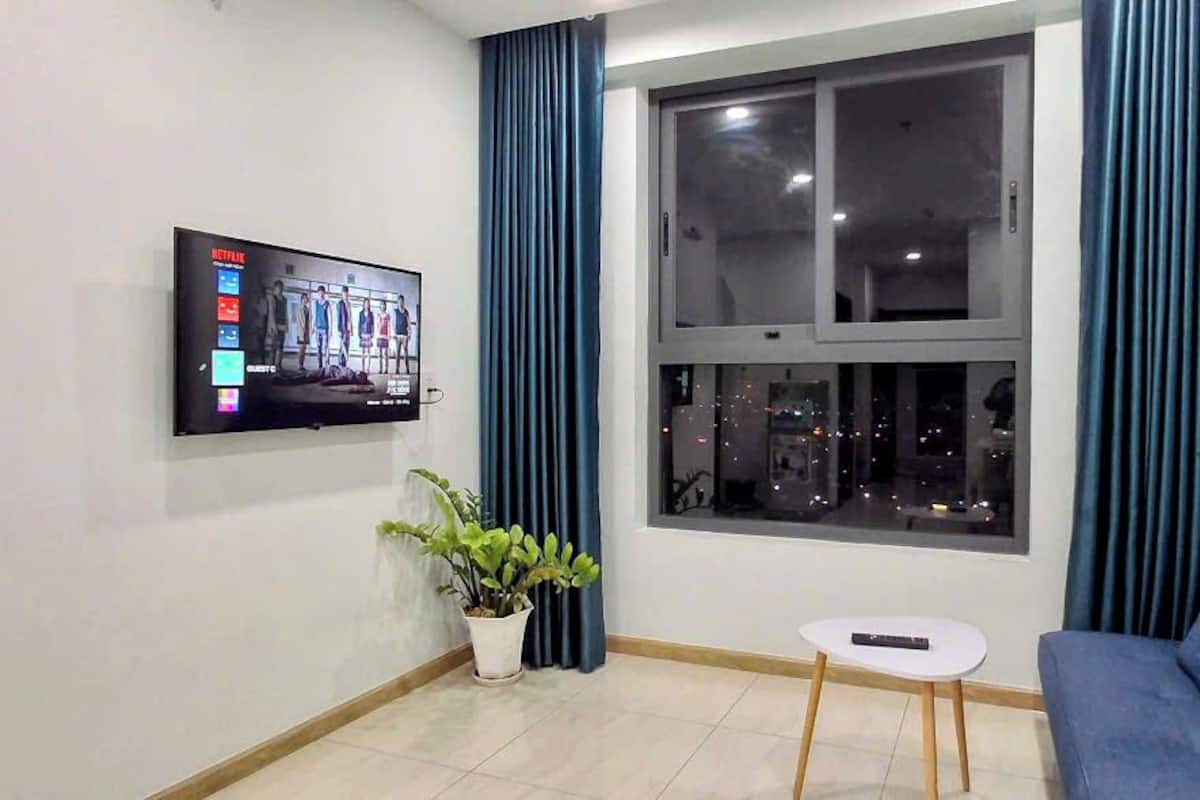 Un salon avec un téléviseur fixé au mur, une plante en pot, une petite table ronde, un canapé bleu et une fenêtre avec des rideaux.