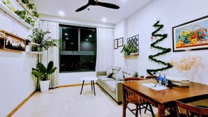 A modern living room with a sofa, dining table, ceiling fan, potted plants, and a large window. - Bcons Garden Luxury Apartment (Ho Chi Minh City)