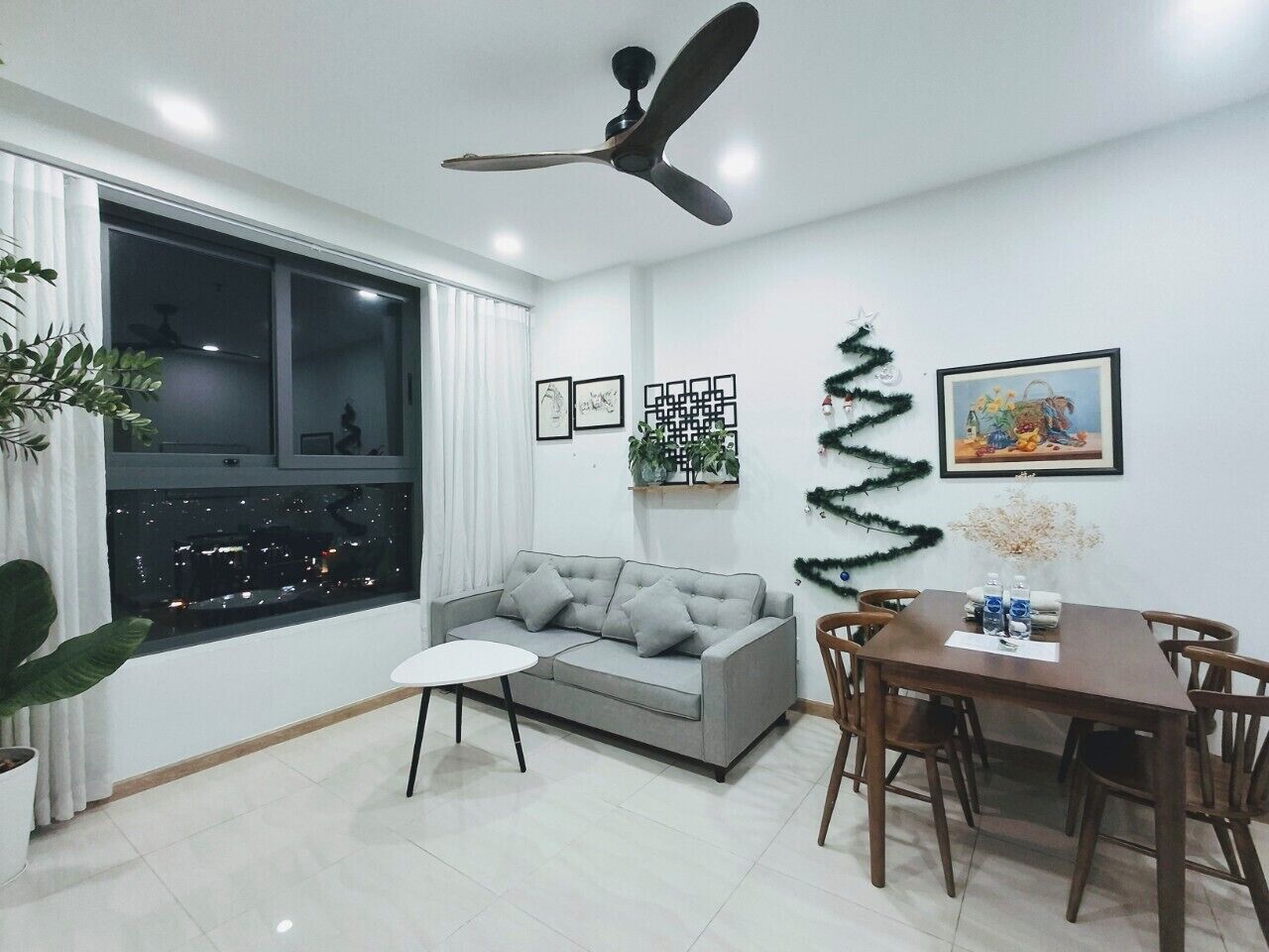Ein modernes Wohnzimmer mit grauem Sofa, einem Esstisch mit Stühlen, einem Deckenventilator und einem großen Fenster mit Blick auf die Stadt.