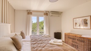 Un dormitori amb un llit gran, una finestra amb cortines, un aparell d'aire condicionat i un tauleig de vimet.
