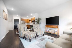 Relax in this stylish living room with a sofa, armchairs, and ottoman, featuring a flat-screen TV and a kitchen area.