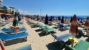 Une plage bordée de rangées de chaises longues et de parasols, un ciel dégagé et des bâtiments en arrière-plan.