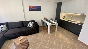 Un espace de vie compact comprenant un canapé de couleur sombre, une petite table ronde en bois, une table à manger blanche avec des chaises, un coin cuisine doté d’appareils électroménagers modernes et un tableau accroché au mur.
