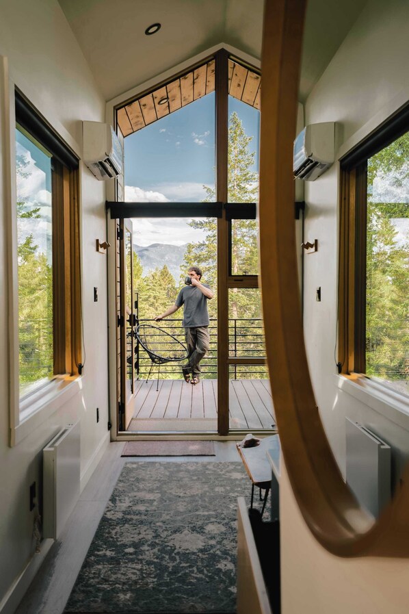 Un interior moderno con un gran ventanal que da a un balcón, una terraza de madera y vistas a montañas.