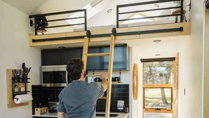 Eine Person erreicht über eine Holzleiter eine erhöht gelegene Küche mit modernen Geräten und einem großen Fenster.
