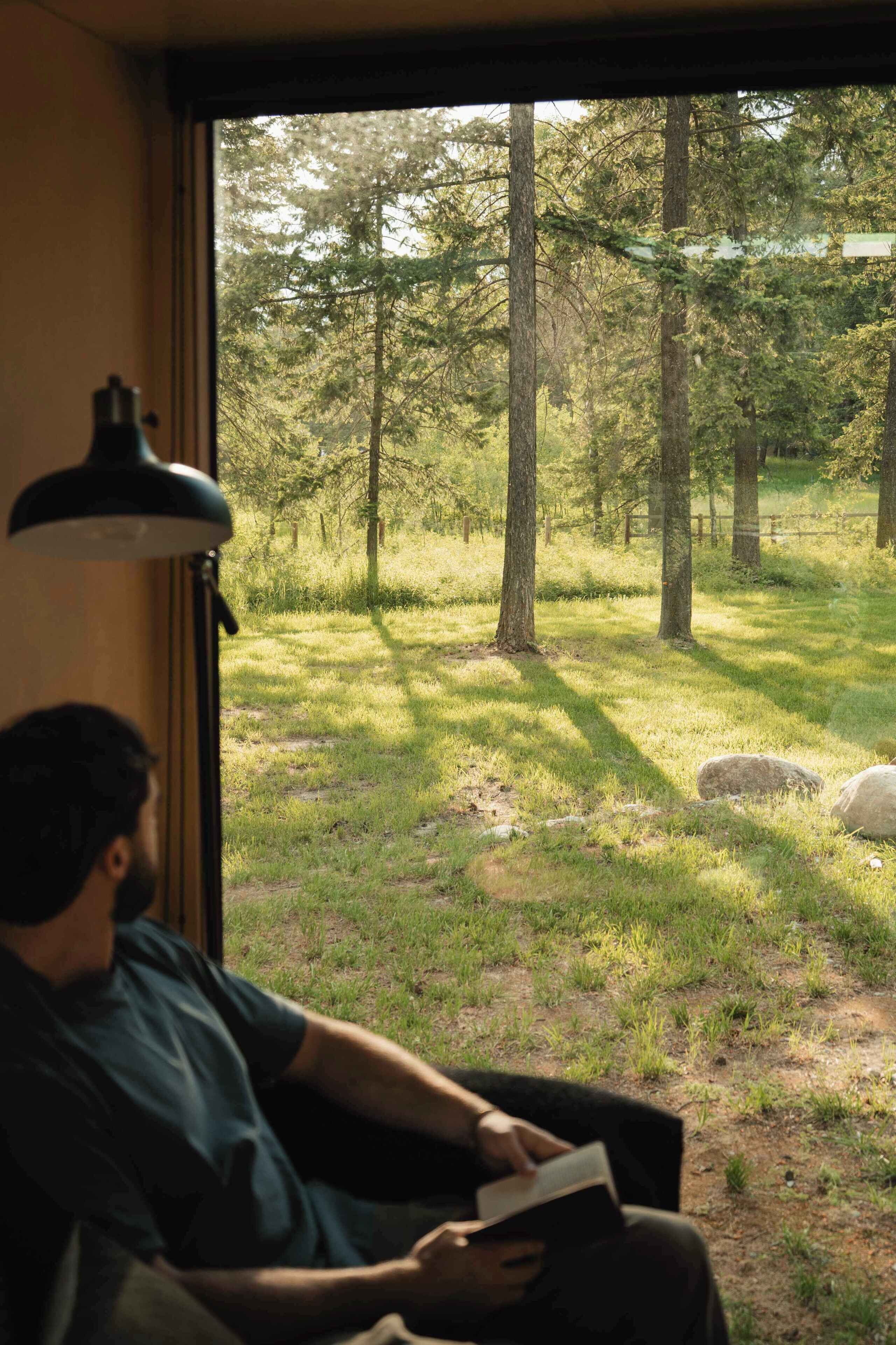 A person sitting on a couch reading a book, with a window view of a forested area.