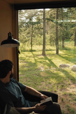 Una persona sentada en un sofá leyendo un libro, con vista a un área boscosa por la ventana.