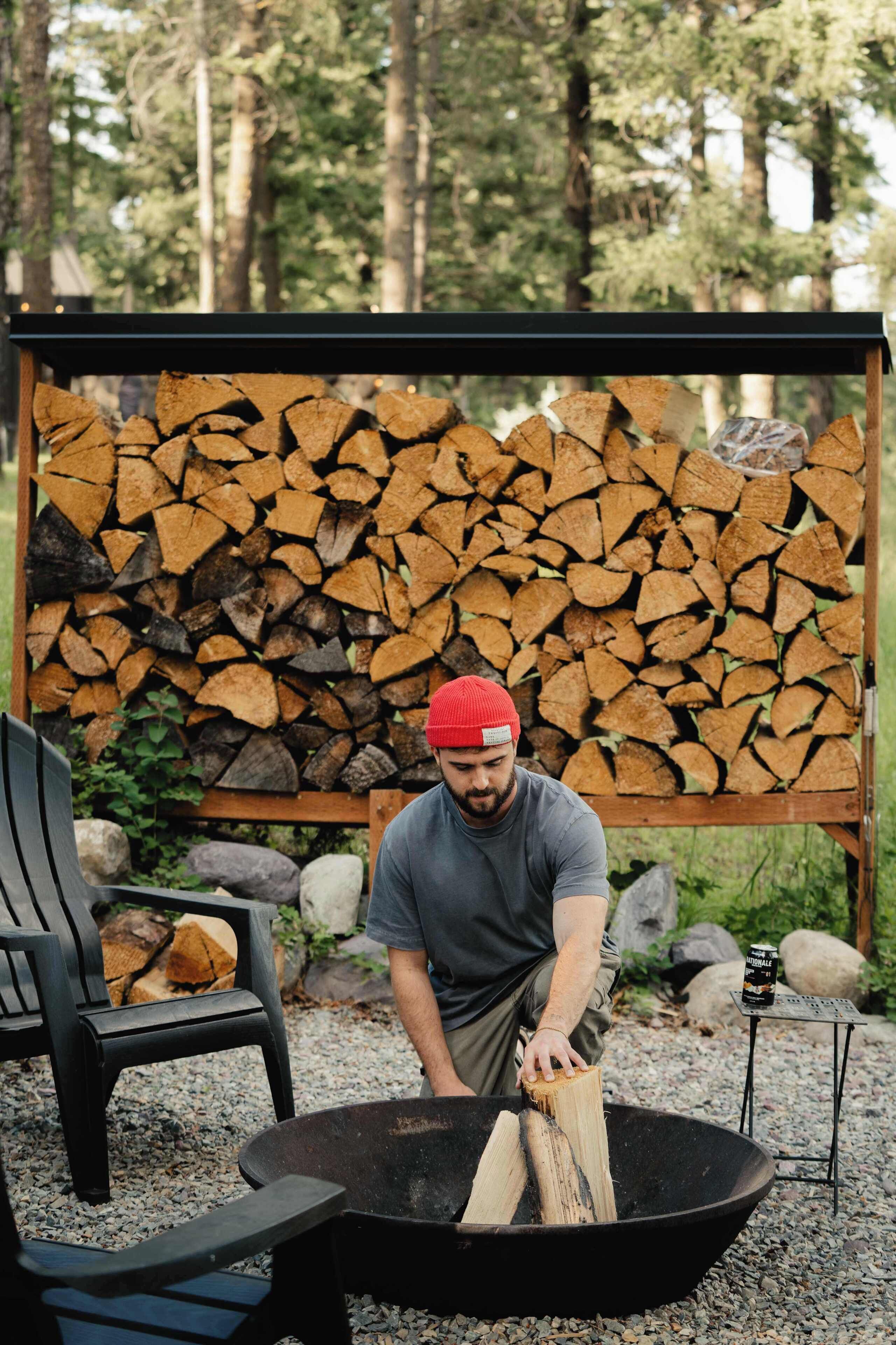 A person sitting by a fire pit with a log, surrounded by a stack of firewood and a chair.
