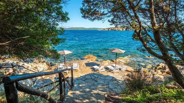 Un sentier côtier avec des marches en pierre menant à une plage aux eaux bleu clair. Des chaises longues et des parasols sont disposés le long du rivage.
