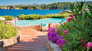 Une piscine bordée de pierres, entourée de verdure et de fleurs, d’où l’on peut admirer la mer et les collines en arrière-plan.