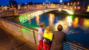 Un pont amb baranes il·luminades sobre un cos d'aigua, amb gent dempeus al passeig proper.