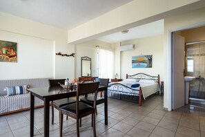 A bedroom with a bed, a bedside table, a window with curtains, a painting on the wall, and a bathroom with a sink and shower.