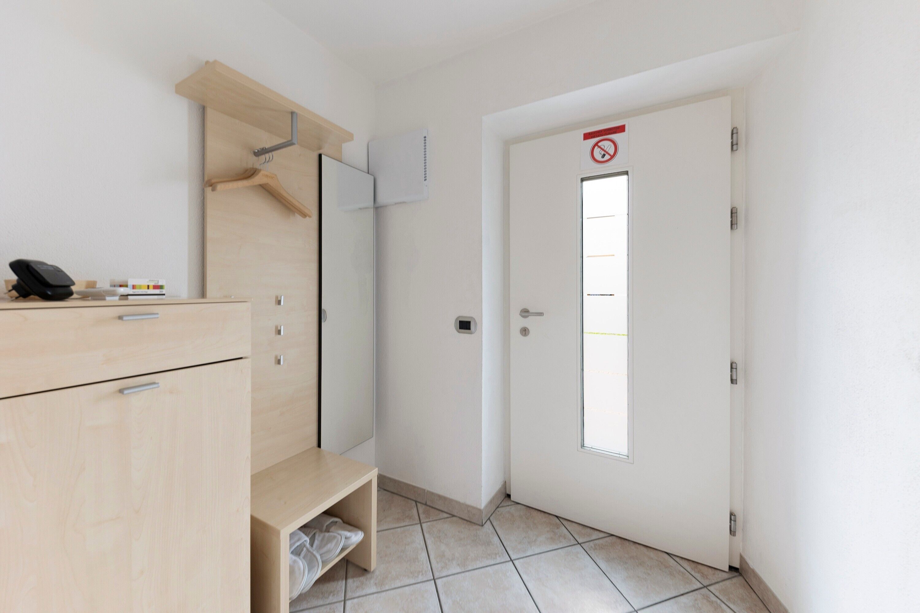 Ein Hotelzimmer mit Holzschrank, einer kleinen Bank, einer Tür mit Rauchverbotsschild und einem Fenster.