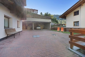 A paved courtyard with a car parked inside a garage, a bench, and a wooden fence. - 'Apartment Bergkristall' with Mountain View, Garden & Wi-Fi (Nova Levante)