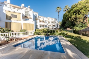 Una piscina con agua azul cristalina, rodeada por un edificio blanco con balcones y un jardín con palmeras.
