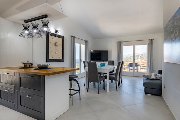 A modern kitchen with a wooden countertop, a dining area with a white table and chairs, and a sofa with a view of the outdoors.