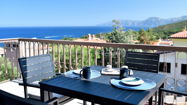 A balcony with a table set for two, overlooking a scenic view of the sea and mountains. - Appartamento Vista Golfo with Sea View, Terrace & Wi-Fi (Cala Gonone)