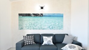 A grey sofa with patterned cushions and a large framed picture of a coastal landscape. - Appartamento Vista Golfo with Sea View, Terrace & Wi-Fi (Cala Gonone)