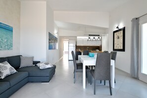 A modern living room with a grey sectional sofa, a dining table with chairs, and a kitchen in the background.