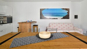 A room with a wooden dining table, a sofa, a kitchen area, and a large beach view painting on the wall. - Appartamento Prato al Mare with Garden, A/C & Wi-Fi (Cala Gonone)