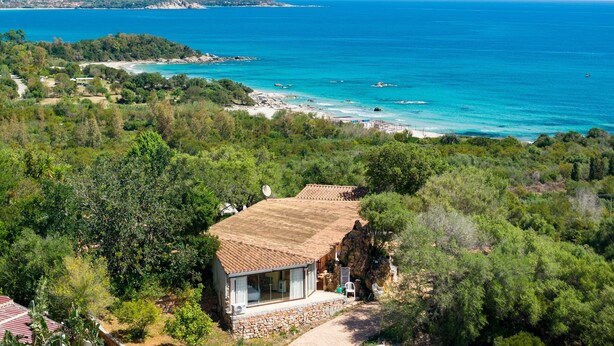 Una casa con techo de tejas, rodeada de abundante vegetación, con vista a una zona costera de aguas azul cristalino y una playa de arena.