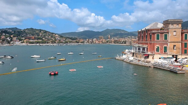 Un pueblo costero con barcos amarrados en el muelle, edificios de fachadas rojas y verdes, y montañas al fondo.