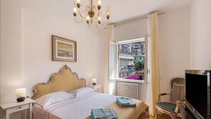 Un dormitorio con cama, mesita de noche, lámpara, televisor, silla y ventana con vistas a edificios.