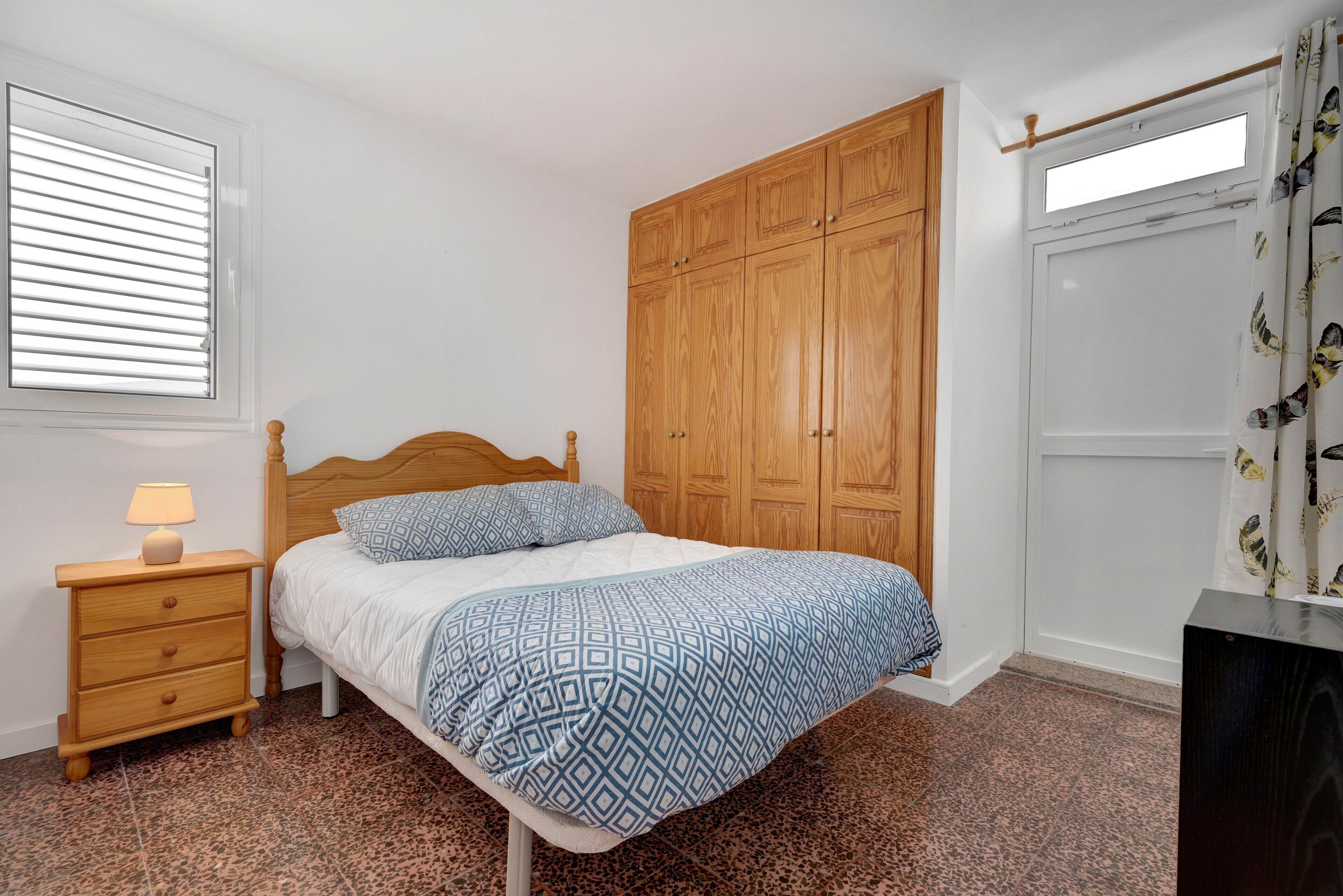 Un dormitorio con cama, armario de madera, mesita de noche con lámpara y alfombra estampada.