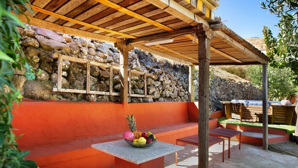 Un espacio de estar al aire libre cubierto con pared de piedra, una pérgola de madera y una mesa con fruta.