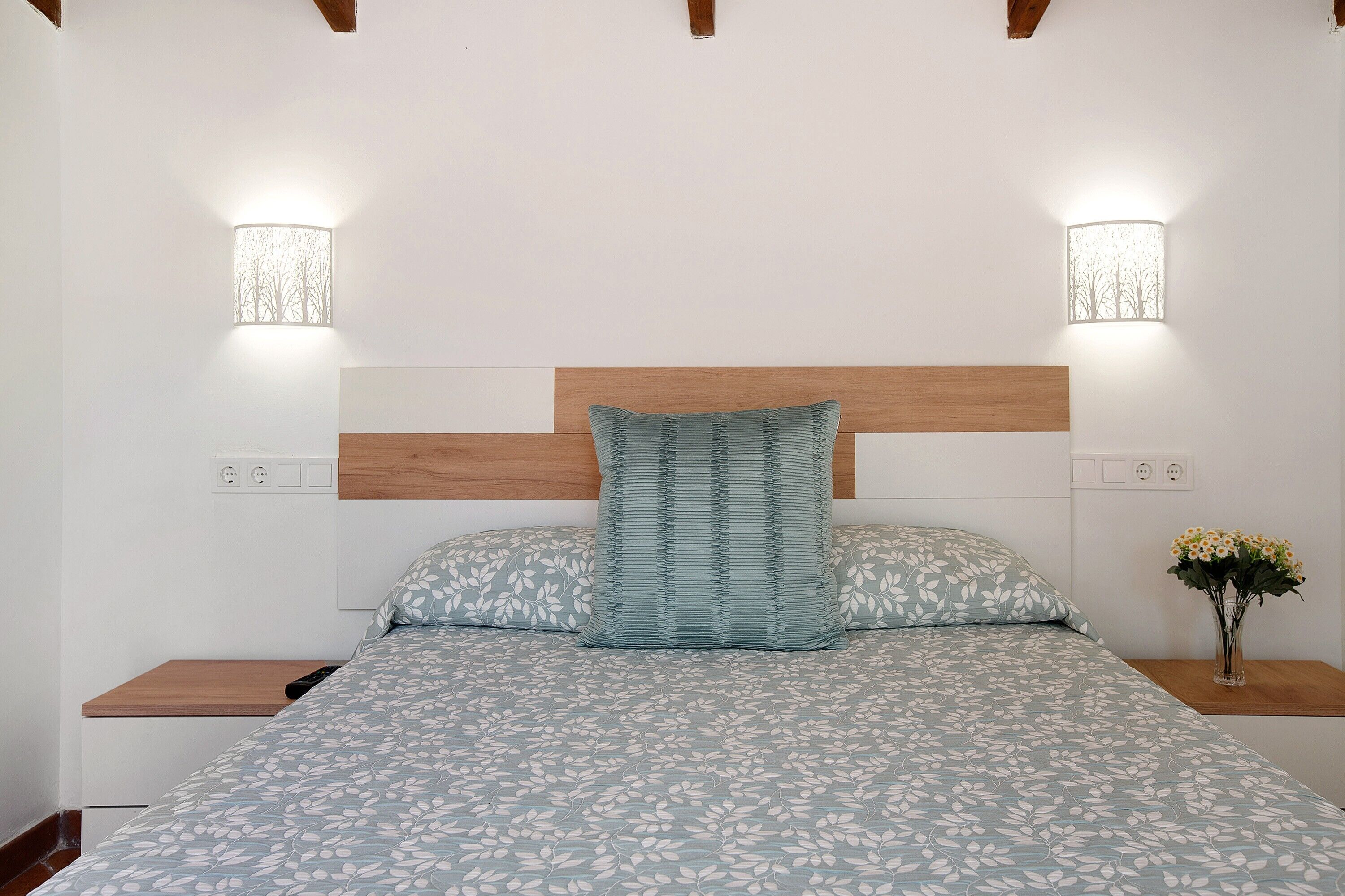 Una cama con colcha estampada, dos almohadas, cabecero de madera, mesitas de noche y luces fijadas en la pared.