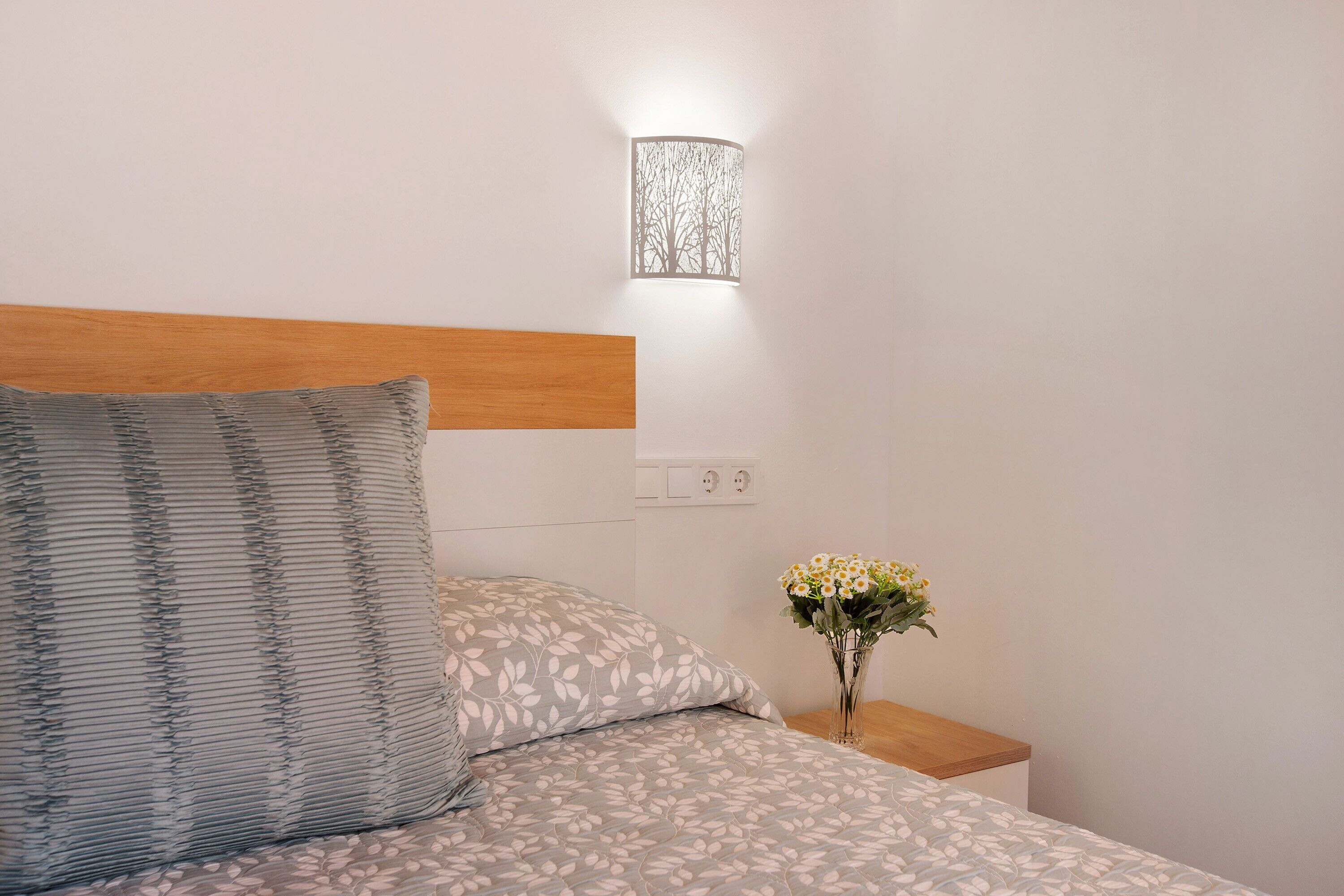 Una cama bien hecha con ropa de cama estampada, una lámpara de noche y un florero con flores sobre una mesita de noche.
