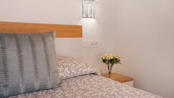 Una cama bien hecha con ropa de cama estampada, una lámpara de noche y un florero con flores sobre una mesita de noche.