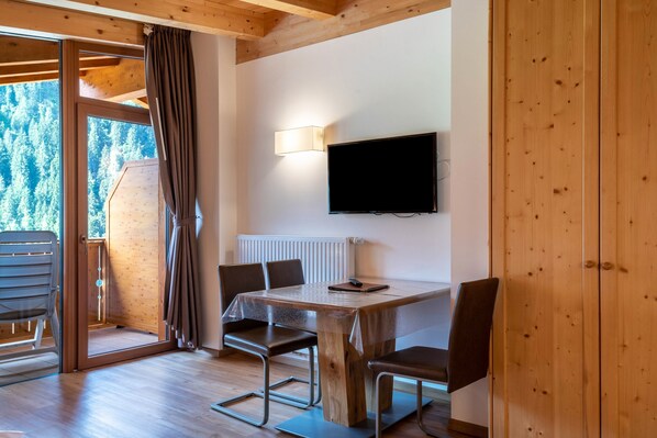 Ein Zimmer mit Holzwänden, einem Tisch, Stühlen, einem Fernseher und Blick auf einen Wald durch eine Schiebetür.