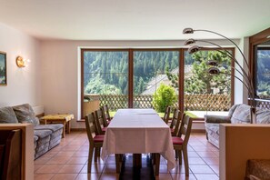 A dining area with a table set for four, chairs, a sofa, a lamp, and a view of a forest through large windows.