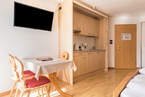 A room with a dining table, chairs, a TV mounted on the wall, a kitchen area, and a door with a map.
